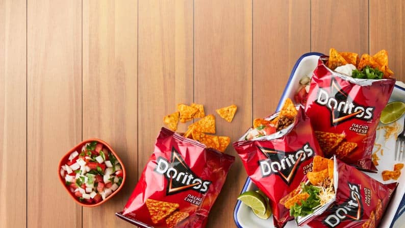 Three walking tacos in bags of Doritos alongside bowls of toppings on a table.