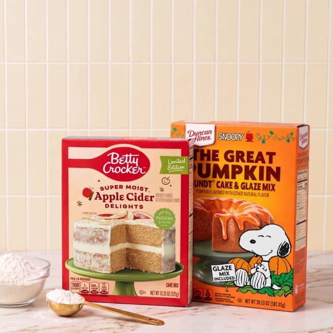 Betty Crocker Apple Cider Delights cake mix and Duncan Hines The Great Pumpkin bundt cake and glaze mix on a kitchen counter with flour and a spoon.
