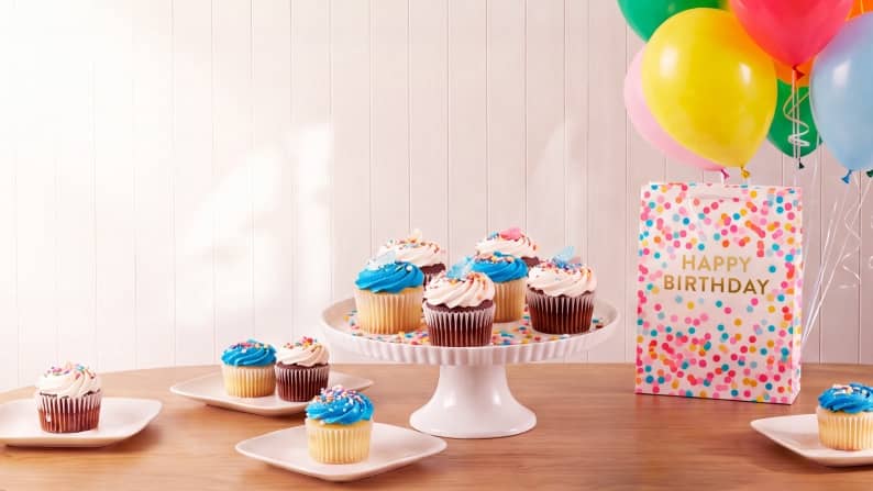 Happy birthday gift bag with balloons beside a cake stand and plates of frosted cupcakes on a wooden table.