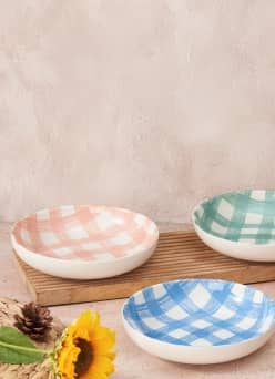 Three plates feature a plaid pattern in pink, blue and green. The plates are on a wooden platter next to a placemat with a sunflower and pinecone.
