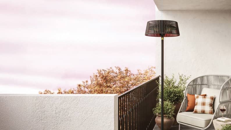 An image of a standing heater on a balcony. A wicker chair overlooks potted green plants and trees with range leaves.