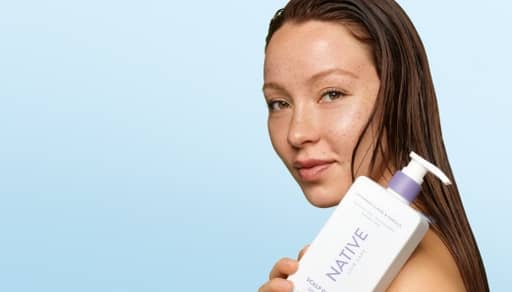 Close-up of a person with wet hair holding a white pump bottle of Native brand hair care against a light blue background.