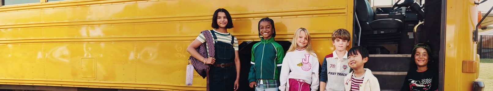 Six children standing in front of school bus with backpacks and wearing back to school outfits.