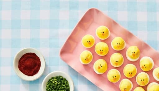 A tray of deviled eggs decorated to look like chicks is pictured with fresh garnish on the side.