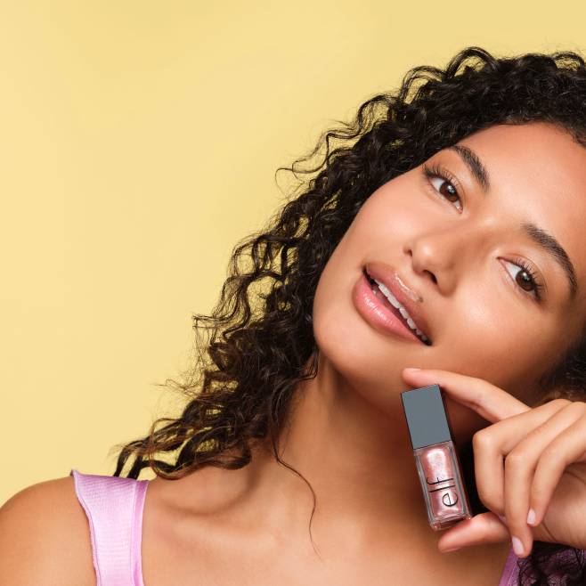 Woman holding pink lip gloss in one hand with text “New makeup, from $9. Shop now” shot against a yellow background.