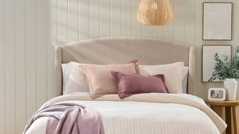 Bed with taupe headboard adorned with mixed bedding of blush and cream duvet, pillows and throws. Nightstand, vase and wall art shown on right.