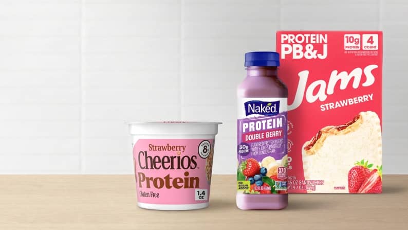 An individual cup of strawberry Cheerios protein cereal, a bottle of Naken protein double berry drink, and strawberry protein PB&J Jams.