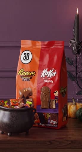 Reeses and KitKat Halloween candy bag with thirty pieces badge beside a cauldron filled with assorted wrapped chocolates and small pumpkins.