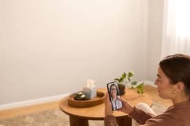 An adult sits in a living room holding a smartphone showing a doctor on a video call near a round coffee table with tissues and a plant.