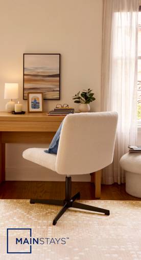A home office is set with a beige swivel chair, natural patterned rug and a wooden desk with decor and a lamp. Sheer white curtains let light in.