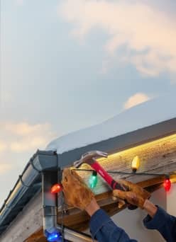 A person hanging colorful Christmas lights on a house roof using a hammer while wearing gloves.