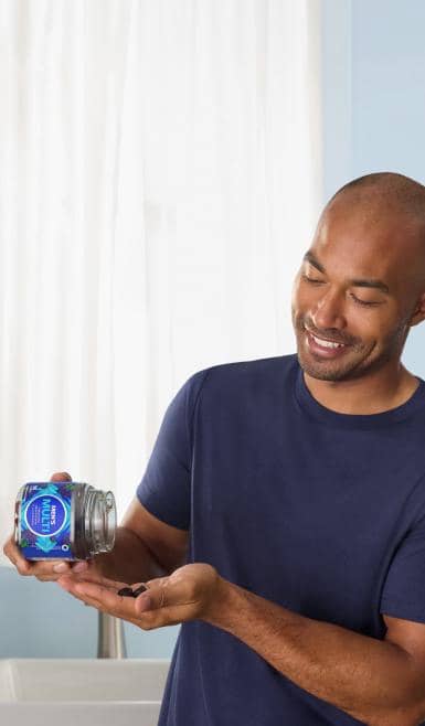 Adult smiles while pouring dark gummies from a clear jar with a blue circular label into their hand indoors.