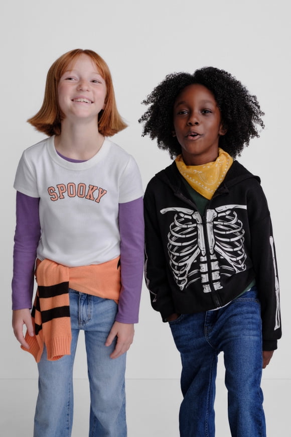 Two children in Halloween outfits, one in a Spooky shirt and one in a skeleton hoodie, stand smiling together.