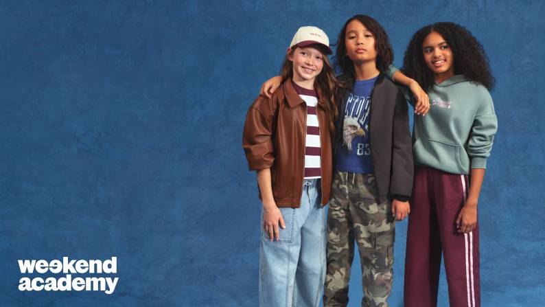 Three tweens stand together against a blue backdrop, arms linked, wearing casual outfits including a cap, jacket, a hoodie, jeans and camo pants.