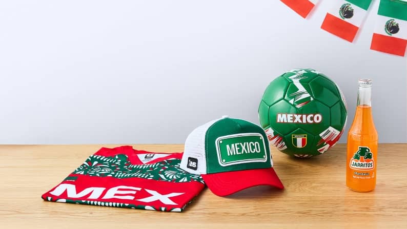 A variety of Team Mexico memorabilia is on a wooden counter—a jersey, a trucker hat, soccer ball, a flag banner and a bottle of Jarritos.
