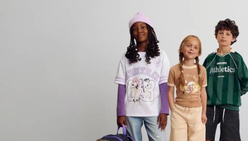 Three children wear a white graphic shirt with a pink beanie, a brown graphic tee and a green striped athletic jersey polo sweatshirt.