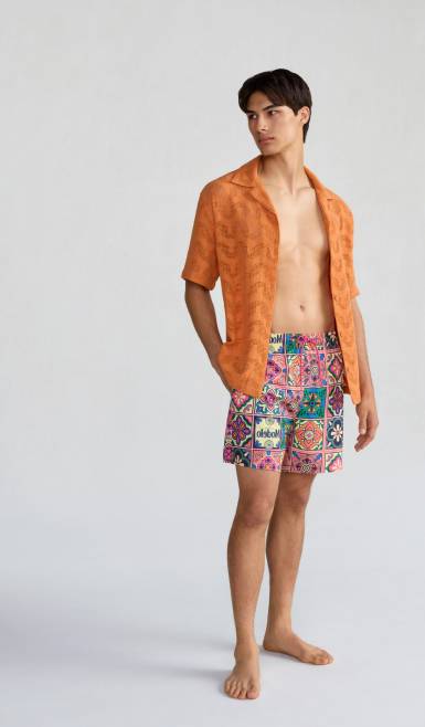 A man wearing an orange shirt and colorful swim shorts is standing.