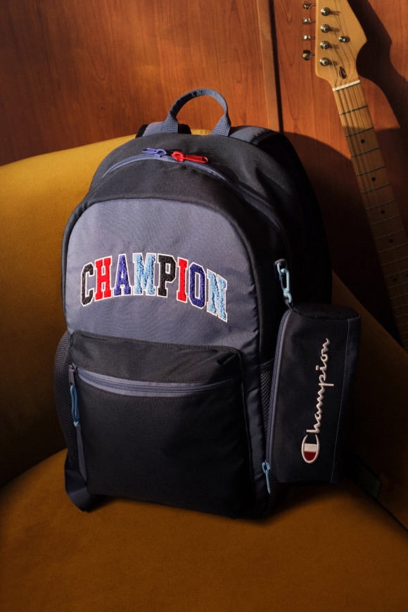 A navy blue Champion backpack is sitting on a yellow chair next to a guitar that is leaning on a wood paneled wall.