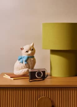 A cat figurine, vintage-style camera and yellow lamp sit on a bedside table in a cozy dorm-inspired setting.
