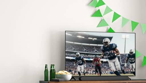 View a media console with a large television showing a football game. Next to the television is a bowl of snacks and drinks.