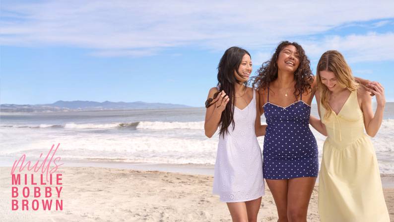Gen Z women in a white mini dress, navy blue polka dot sundress, and yellow midi dress on a beach; trendy coastal summer aesthetic.