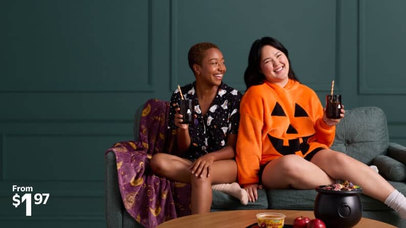 Image shows two women in Halloween loungewear, sitting on a couch with drinks, a blanket and plate of food. From one dollar and ninety-seven cents.