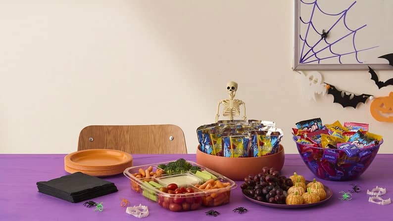 A vegetable and dip tray, a bowl of Capri-Sun pouches, a tray of grapes and mandarin oranges, and a bowl of Nerds, M&M’s, Trolli & other candies.