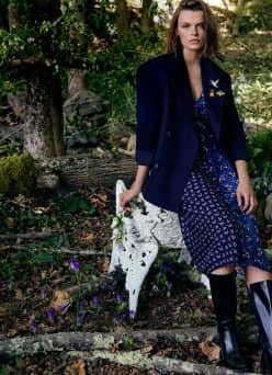 Woman in a navy blazer over a patterned dress and knee-high boots, posing in a wooded garden.