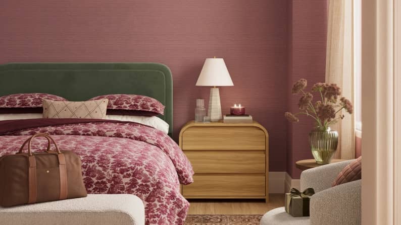 Bedroom with green headboard, mauve patterned bedding, wooden nightstand with lamp and candle, bench with travel bag and armchair.