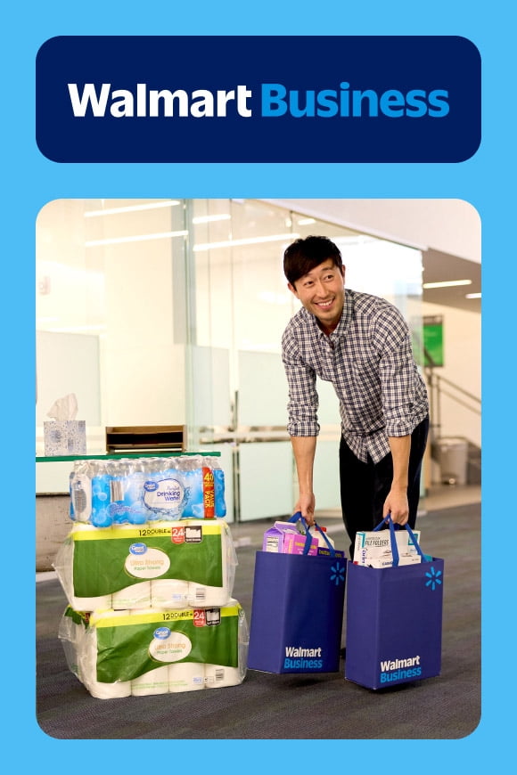 A smiling man in an office sets down two grocery bags next to a stack of paper towels. The Walmart Business logo is at the top.