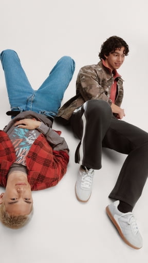 Two men wearing a red plaid shirt with jeans and a bomber jacket over a red tee with black pants and white sneakers.