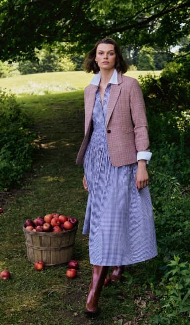 Woman in a blue gingham dress, checked blazer and brown boots in an apple orchard.