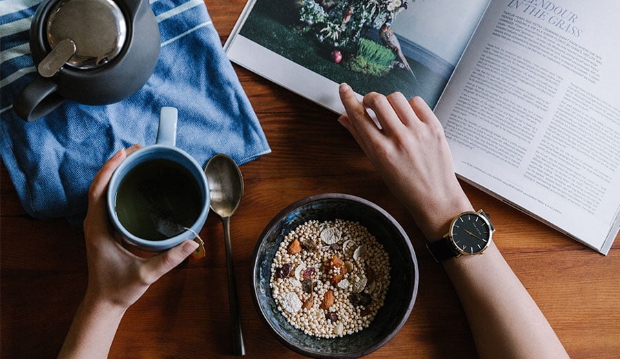 Top down view of some eating breakfast, their left hand is holing a mug of tea with a tea pot next to them, their right hand is flipping the page of a book. There's a bowl of oatmeal in between their arms.