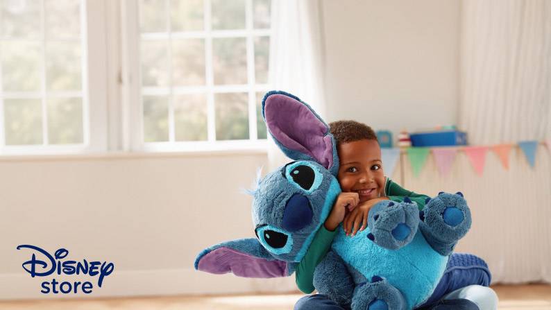 Disney Store. A smiling child hugs a large blue Stitch plush toy while sitting indoors.