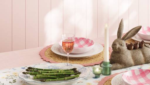 A table with bunny and bird figures, white and pink plates, a plate of asparagus, a glass of wine, a flower tablecloth, placemats and candles.