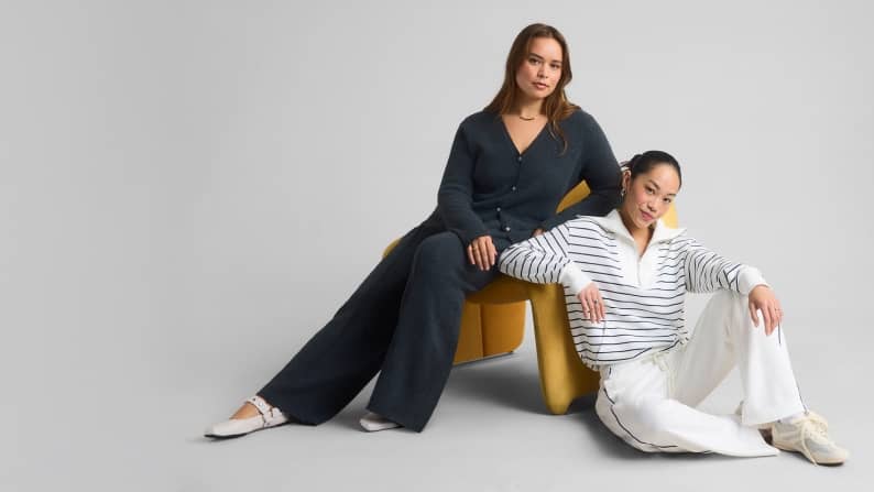 A woman wearing dark gray loungewear seated in a chair next to a woman wearing white loungewear seated on the ground in front of a gray background.