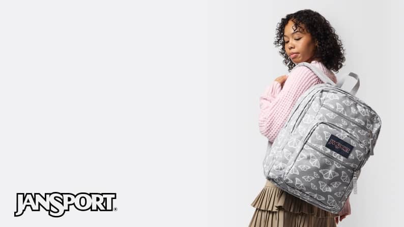 Young adult woman in a pink top and tan skirt with a grey patterned JanSport backpack on white background.