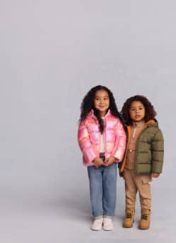 A toddler girl and boy are shown. She wears a pink shiny puffer jacket. He wears a green puffer jacket over a camel quarter zip pullover and joggers