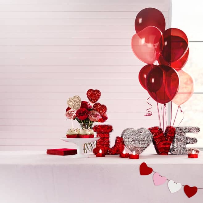 A party table is decorated with red balloons, red napkins, a red heart table bouquet, red heart banner, and sparkly letters that spell love.