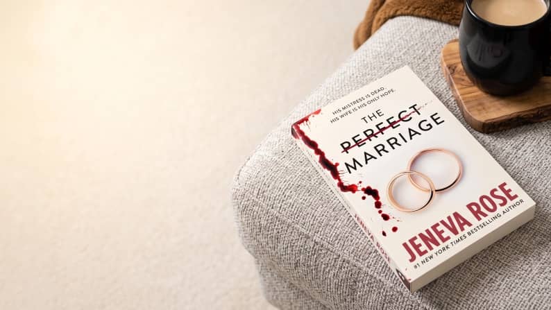 The Perfect Marriage by Jeneva Rose paperback with gold rings and blood splatter on a gray ottoman beside a black mug on a wooden coaster.