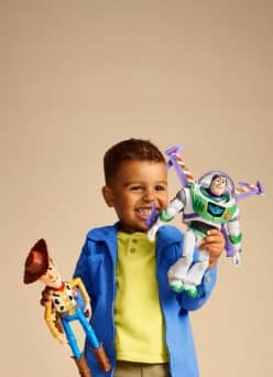 Smiling child in a blue jacket holding Woody and Buzz Lightyear action figures against a plain background.