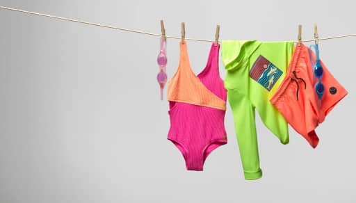 A pair of pink goggles, a colorful one-piece, a neon rashguard and a pair of orange swim shorts are on a clothesline.
