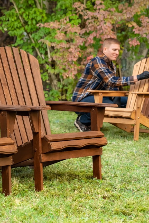 An Adirondack chair in the foreground with a man restaining another chair in the background
