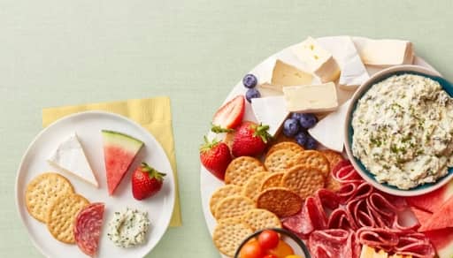 A charcuterie plate is shown with dip, cheese, meat, crackers and fruit next to a plate with crackers, cheese & fruit.