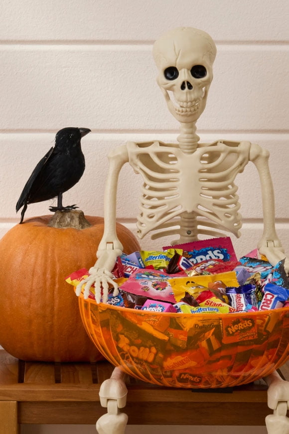 Plastic skeleton holds a large orange bowl filled with assorted wrapped Halloween candy beside a pumpkin and a black bird.