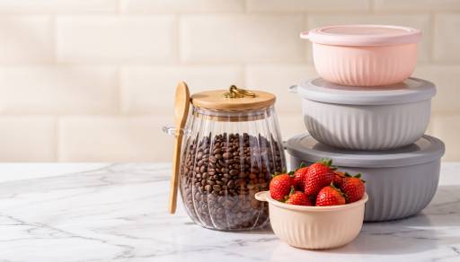 Glass jar of coffee beans with wooden lid and spoon beside bowls of fresh strawberries and stacked pastel food storage containers on a counter.