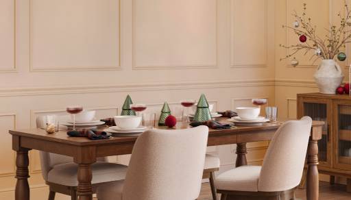 Holiday dining room with wooden table set with small Christmas trees, red ornaments and glasses of red drink, surrounded by upholstered chairs.
