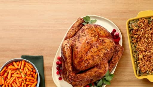 A dish of green bean casserole is shown next to a roasted turkey on a platter garnished with cranberries and a bowl of baby carrots and herbs.