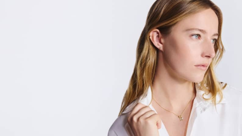 A woman wearing a white blouse and delicate gold necklace, looking to the side against a light background.