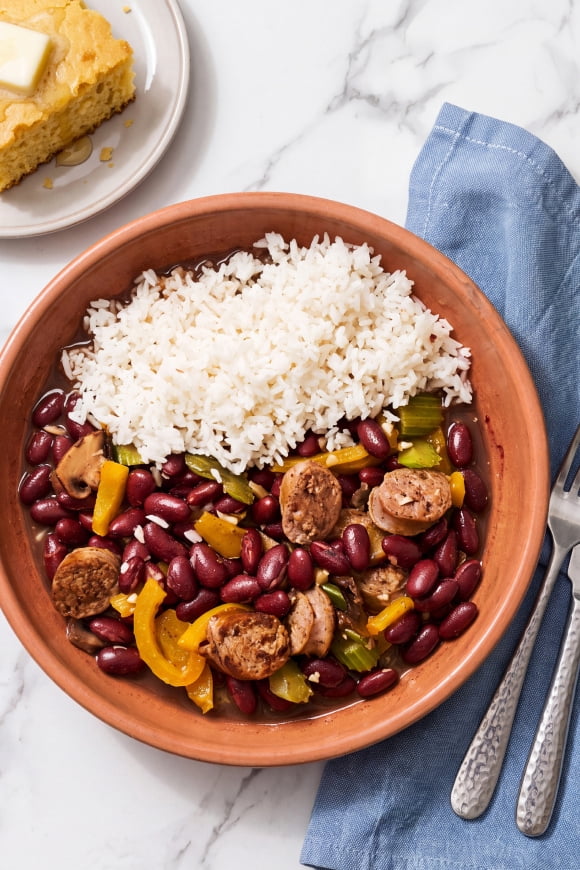 Clay bowl of white rice with red beans, sausage and vegetables on marble beside blue napkin, spoons and cornbread with butter.